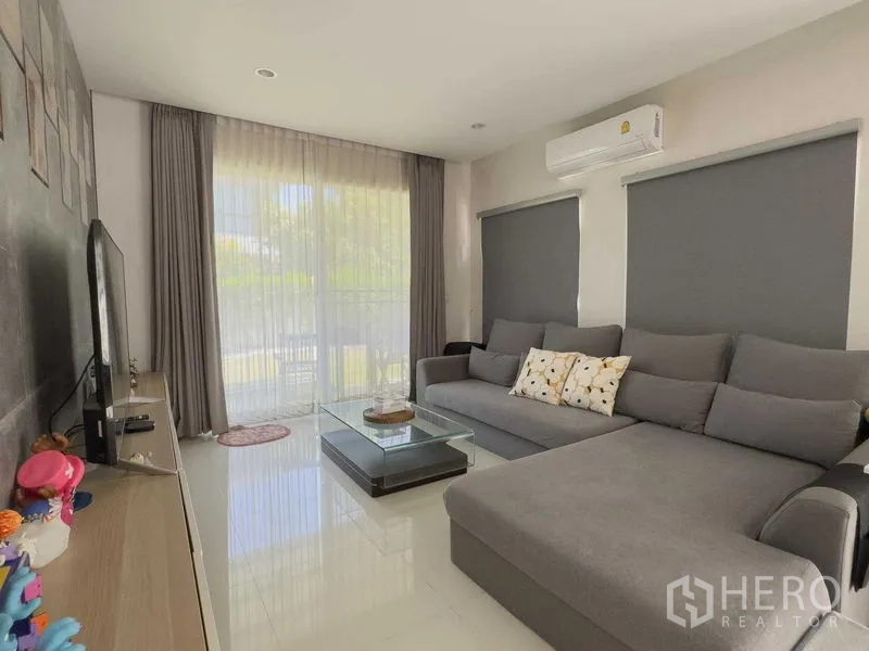 Detached House for rent or sale in Bang Khen, Bangkok - Cozy living room with L-shaped gray sofa, TV, large windows and air conditioning.