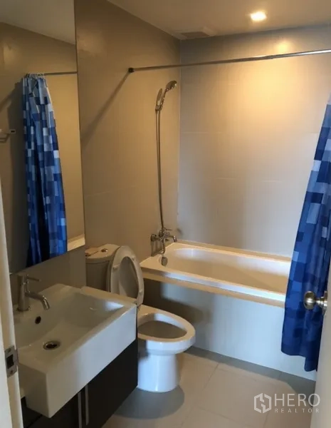 Condominium for rent in Khlong Toei, Bangkok - Bathroom with bathtub and shower, white sink vanity, toilet and blue shower curtain.
