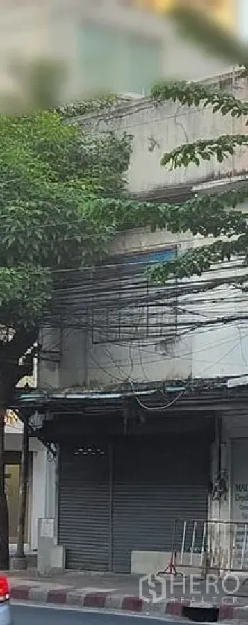Shophouse for rent in Watthana, Bangkok - Street-front two-storey shophouse exterior with closed metal roller shutter, sidewalk curb, leafy tree and overhead cables in urban Ekkamai, Bangkok.
