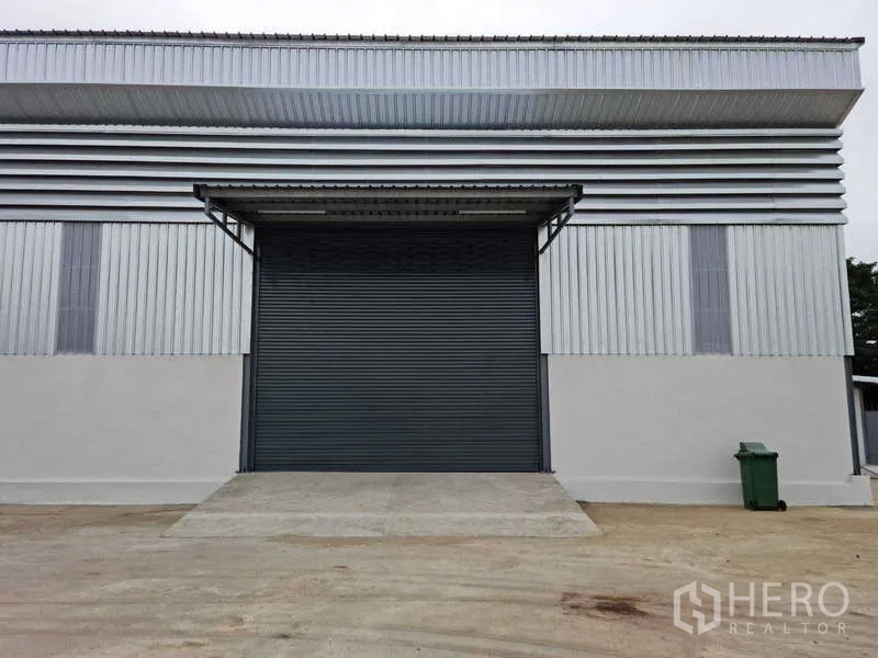 Warehouse for rent in Bang Bua Thong, Nonthaburi - Modern warehouse exterior with large roller shutter, metal cladding and covered loading bay on a wide concrete apron.
