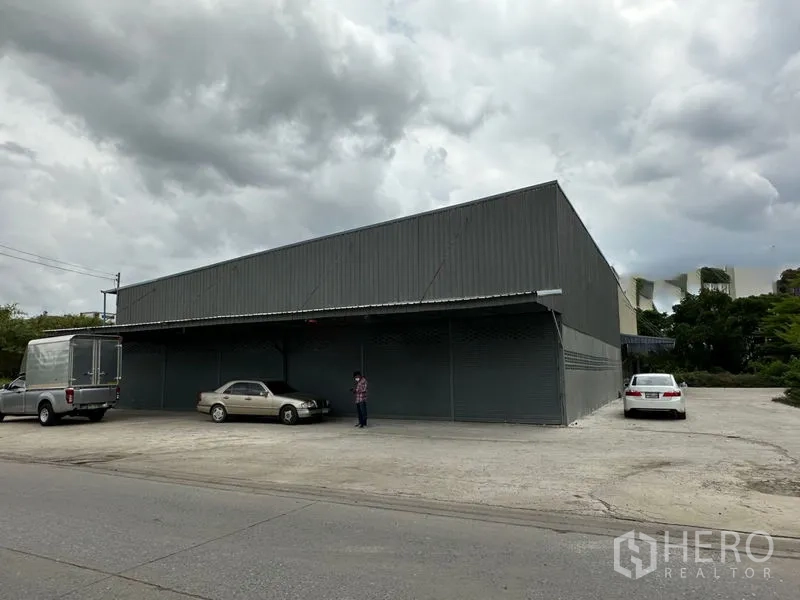 Warehouse for rent in Thanyaburi, Pathum Thani - Front exterior of a gray metal warehouse with wide concrete forecourt, roadside access and roller shutter doors.