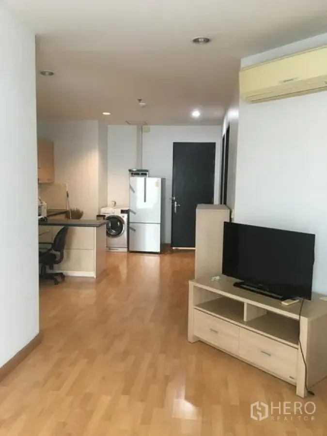 Condominium for rent in Khlong Toei, Bangkok - Open-plan living area with light wood floors, TV console, wall air conditioner, and kitchen with fridge and washing machine.