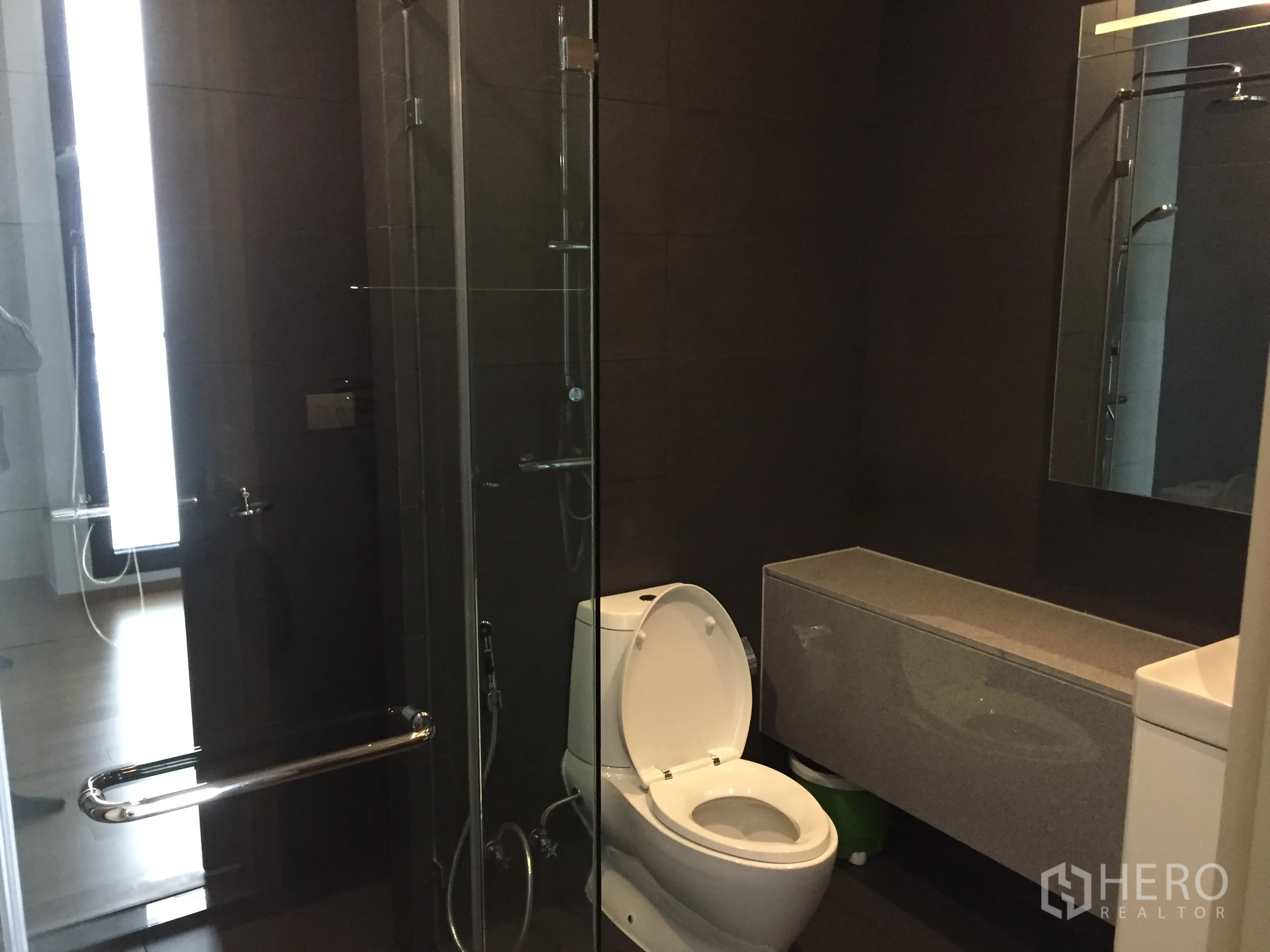 Condominium for rent in Pathum Wan, Bangkok - Modern bathroom with glass shower enclosure, floating vanity and toilet in dark tiles.