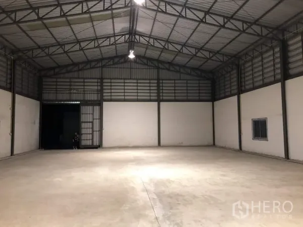 Warehouse for rent in Lam Luk Ka, Pathum Thani - Clear‑span warehouse looking toward the loading door with ceiling lights and ventilated upper walls.