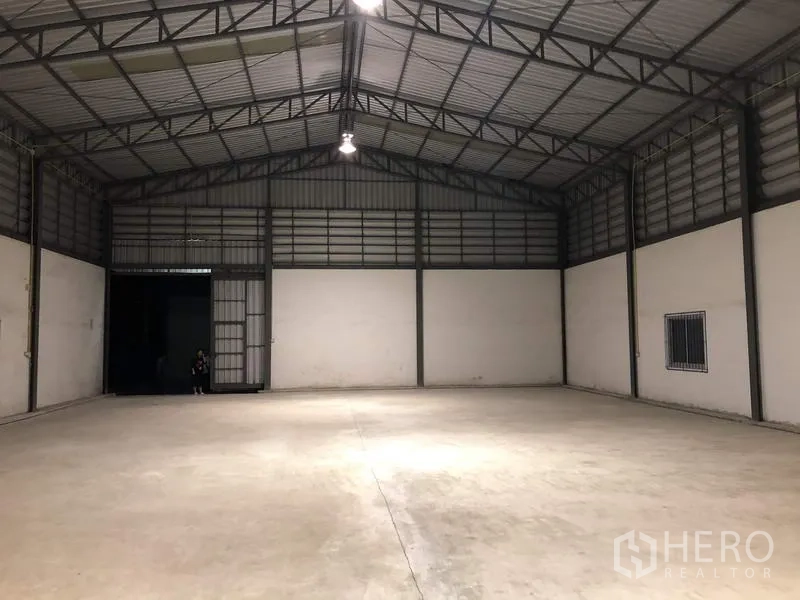 Warehouse for rent in Lam Luk Ka, Pathum Thani - Clear‑span warehouse looking toward the loading door with ceiling lights and ventilated upper walls.