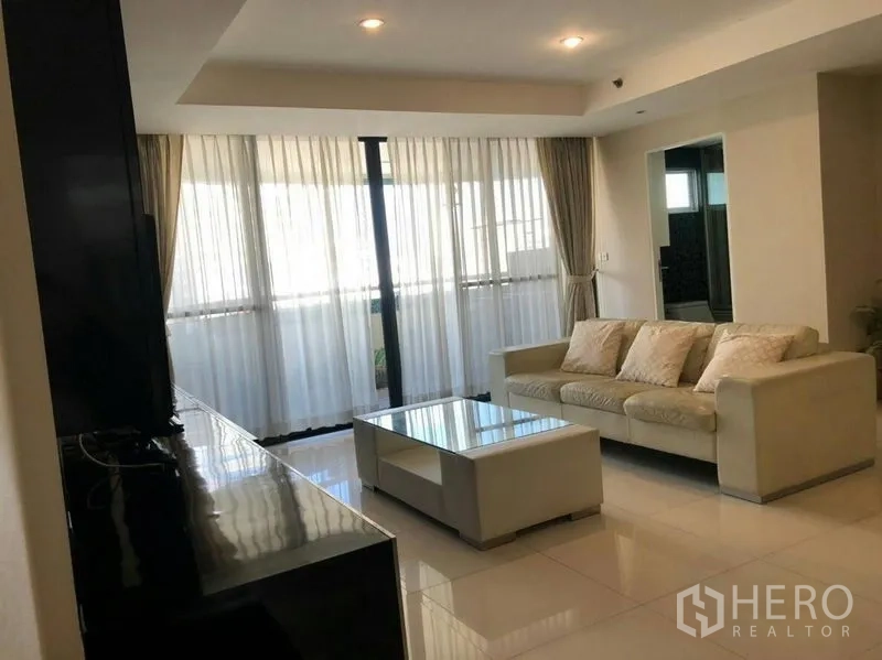 Condominium for rent or sale in Watthana, Bangkok - Lounge area with sofa and glass coffee table next to a wall of windows and curtains.