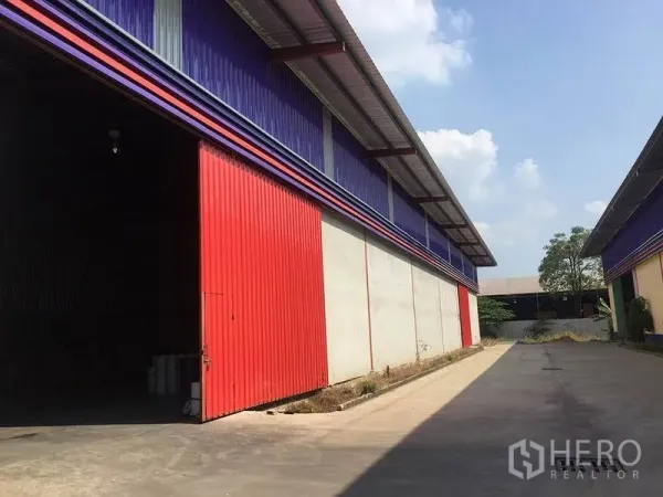 Warehouse for rent in Sai Noi, Nonthaburi - Side view of warehouse showing a red sliding door and a wide truck-friendly concrete lane between buildings.