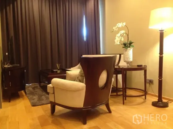 Condominium for rent in Khlong Toei, Bangkok - Cozy lounge corner with upholstered armchair, round side tables and tall floor lamp against dark curtains.