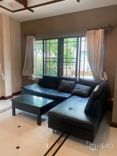 Detached House for rent or sale in Mueang Nonthaburi, Nonthaburi - Bright living room with L-shaped black leather sofa, glass coffee table and large windows.