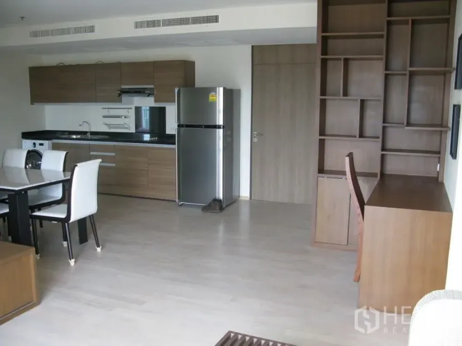 Condominium for rent in Khlong Toei, Bangkok - Open-plan living area with wood cabinetry kitchen, stainless fridge, built-in shelving and desk.