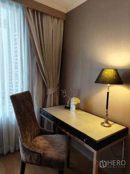 Condominium for rent or sale in Pathum Wan, Bangkok - Compact work corner with upholstered chair, slim writing desk, and table lamp next to curtains.