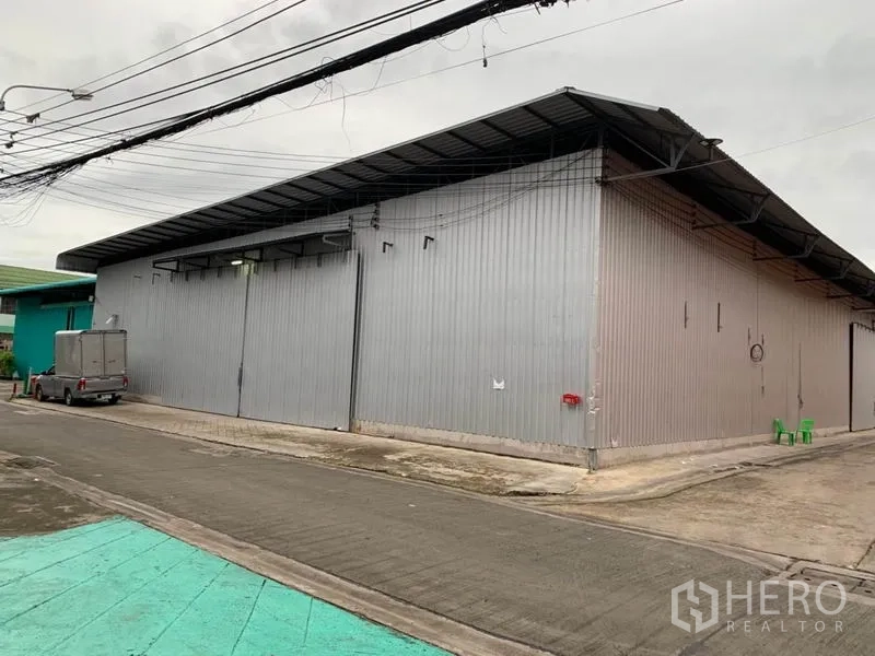 Warehouse for rent in Bang Kapi, Bangkok - Metal-clad warehouse exterior with wide sliding doors and truck access on a paved lane in Bang Kapi, Bangkok.