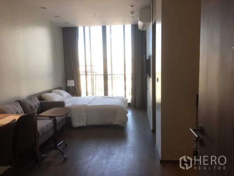 Condominium for rent in Khlong Toei, Bangkok - Bright room with floor-to-ceiling windows, balcony rail, bed, sofa and wall-mounted TV.