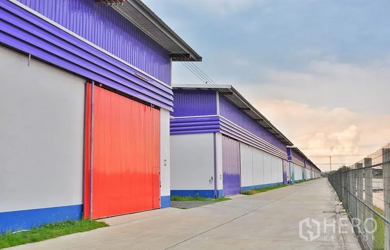 Warehouse for rent in Pathum Thani - Row of modern purple-and-white warehouse units with large red sliding doors and a wide truck-friendly driveway.