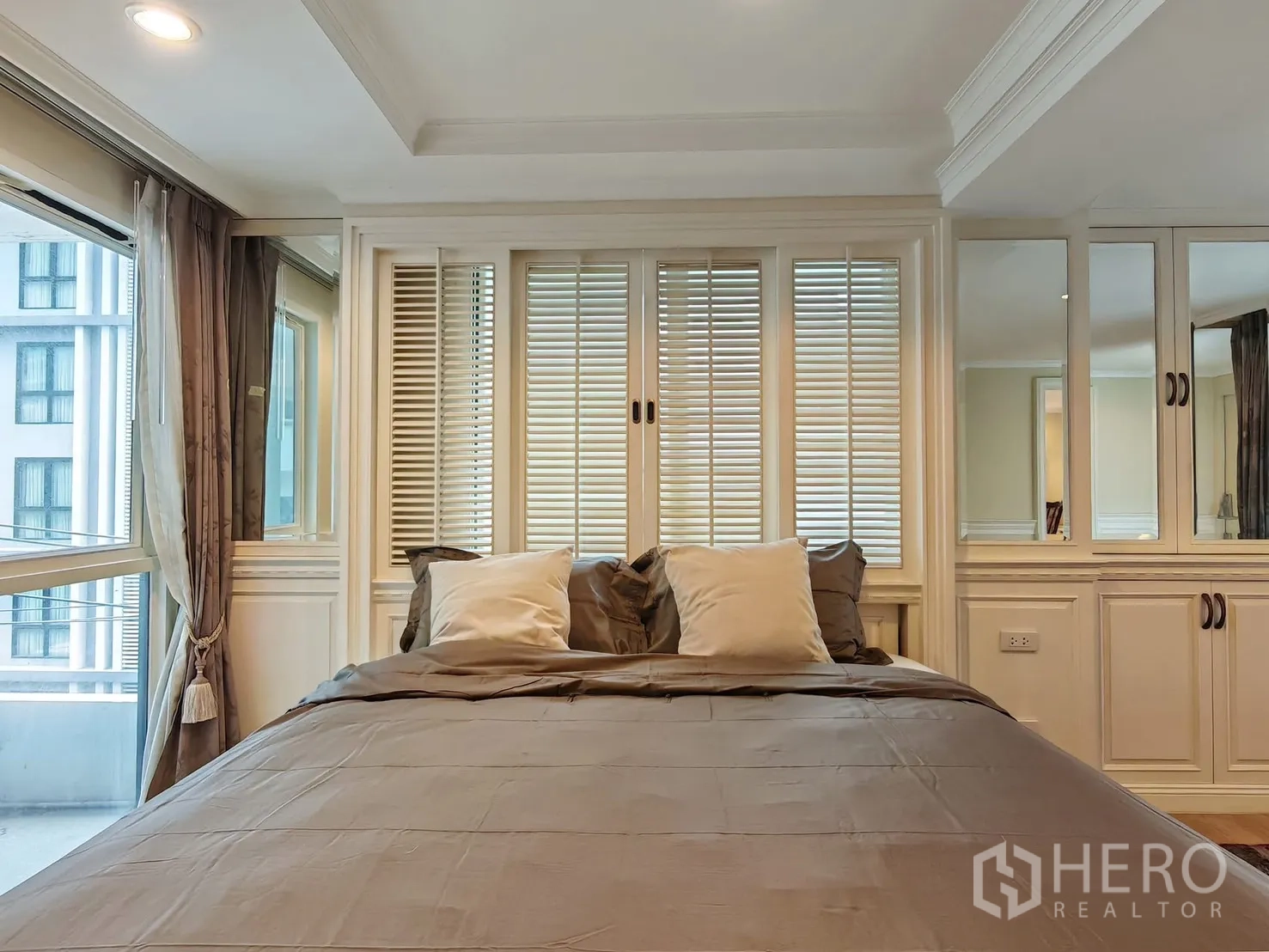 Condominium for rent in Pathum Wan, Bangkok - Bed beside large windows with louvered panels and mirrored cabinets.