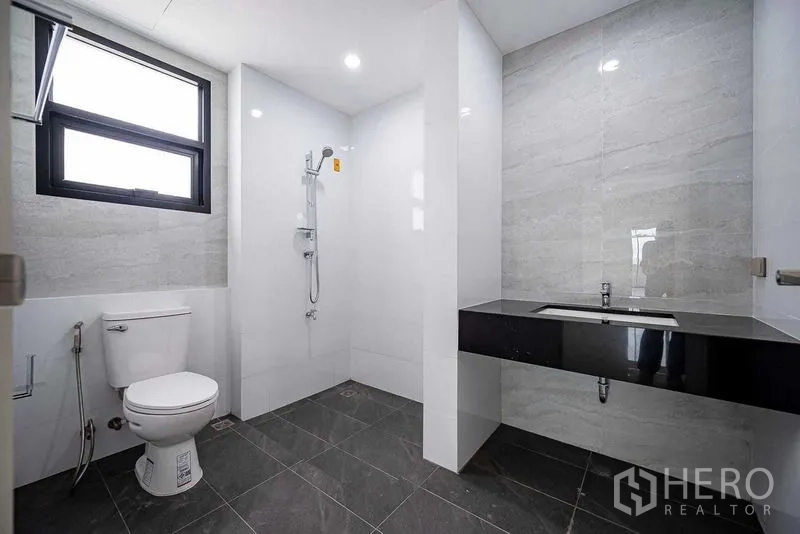 Home Office for rent in Suan Luang, Bangkok - Bathroom with walk-in shower, toilet, and wide vanity set on dark gray floor tiles.