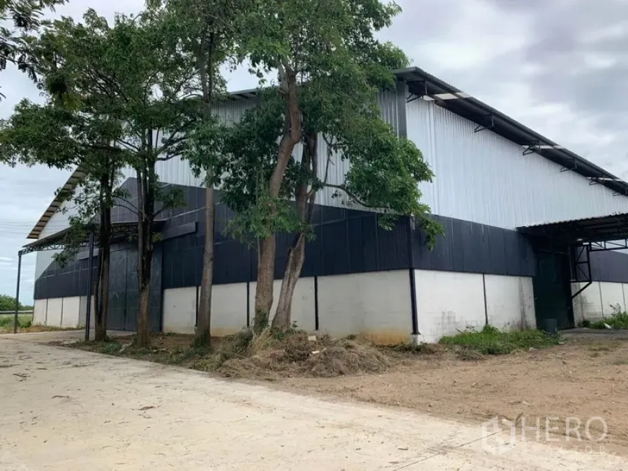 Factory for rent in Chon Buri - Large steel-clad factory exterior with mature trees and a concrete driveway under a cloudy sky.