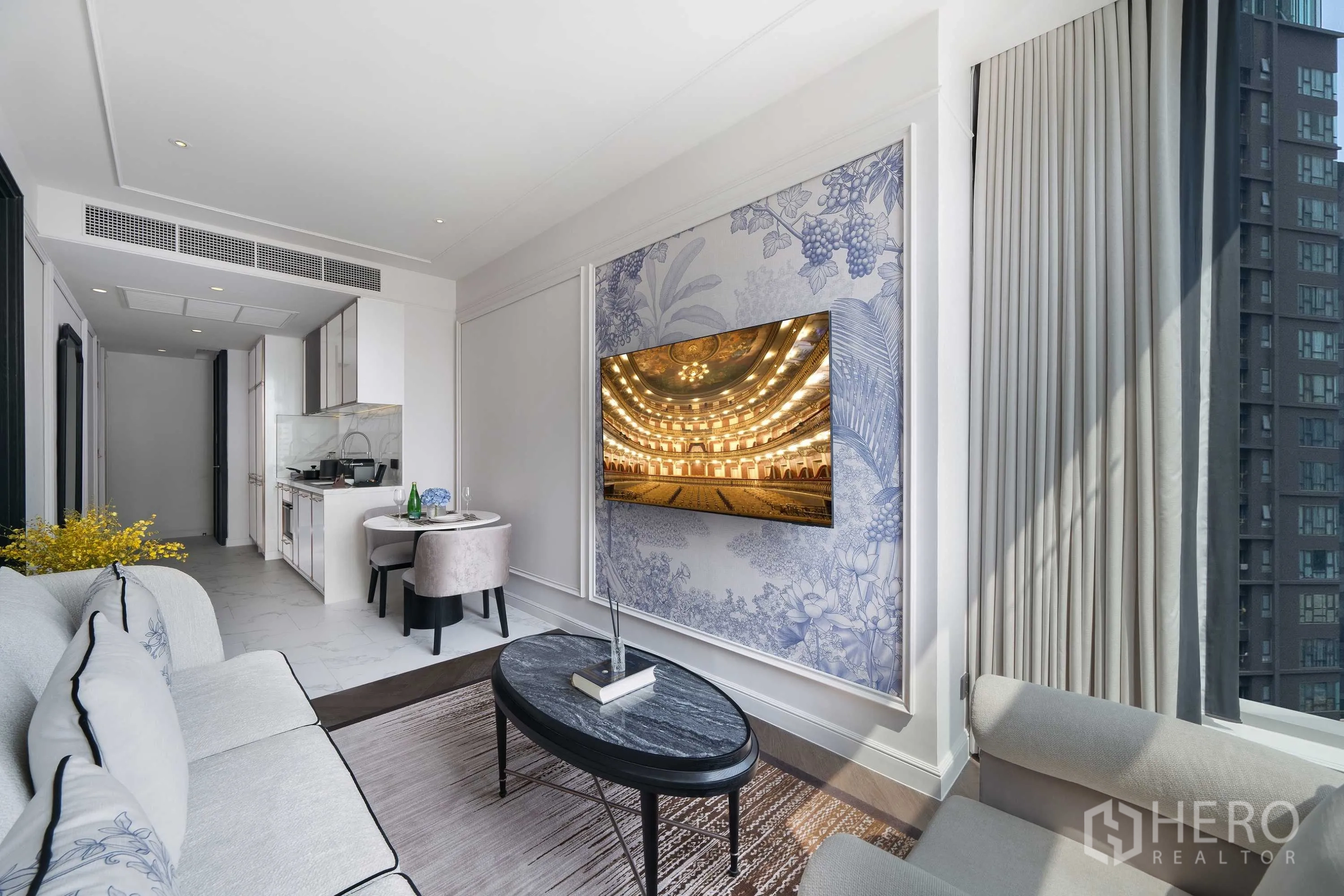Condominium for rent in Khlong Toei, Bangkok - Living area with a mounted TV on a decorative wall, oval coffee table, tall curtains, and urban skyline outside.