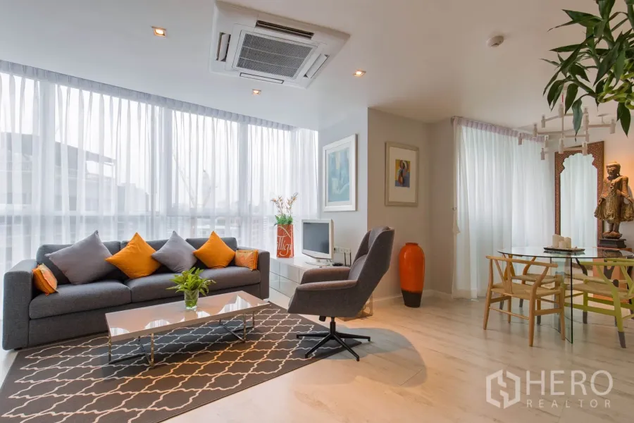 Condominium for rent in Pathum Wan, Bangkok - Bright open-plan living and dining area with floor-to-ceiling windows, gray sofa with orange cushions, armchair, and light wood floors.