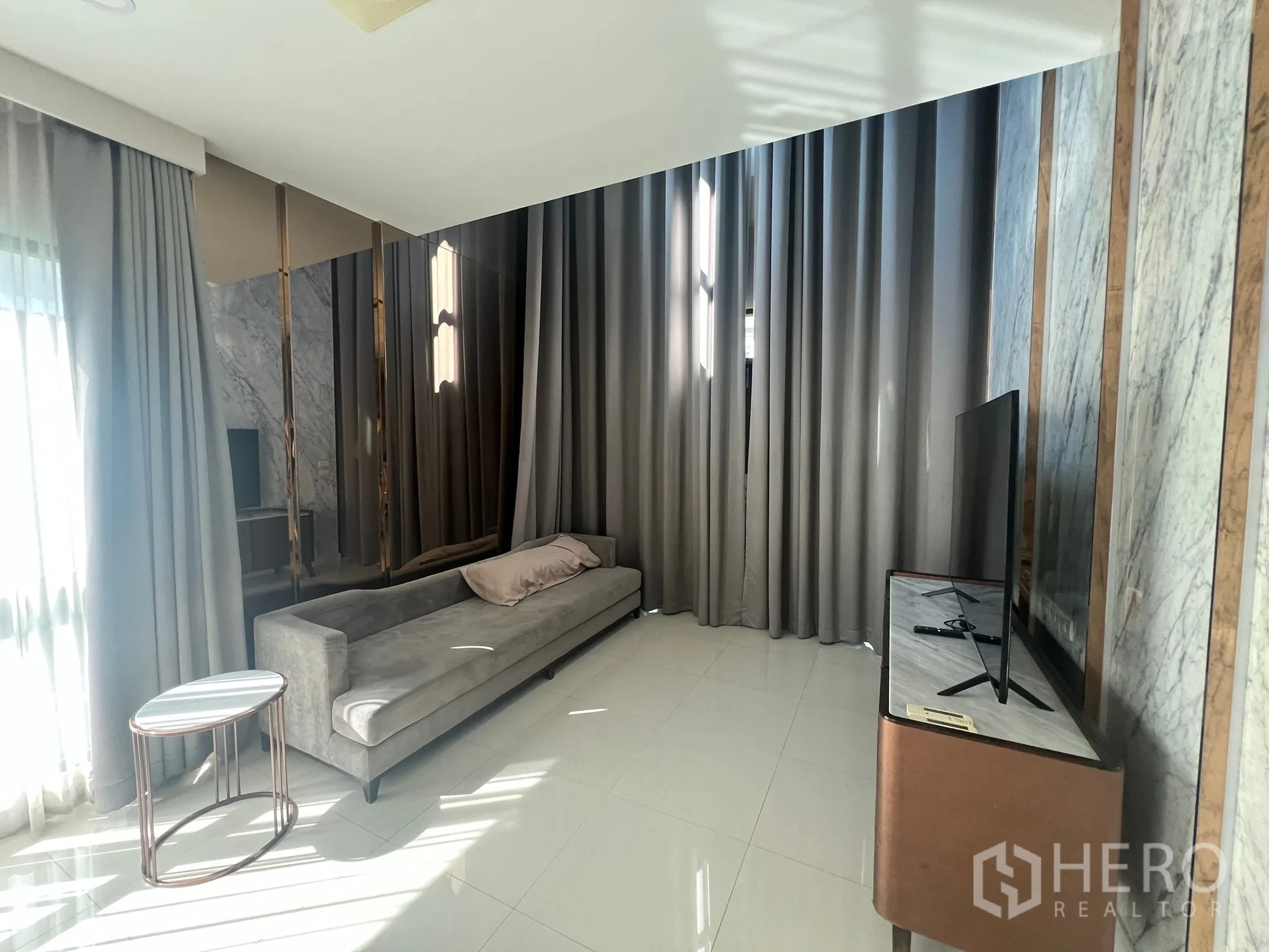 Detached House for rent or sale in Prawet, Bangkok - Bright lounge with a long gray sofa, floor-to-ceiling curtains and a TV console.