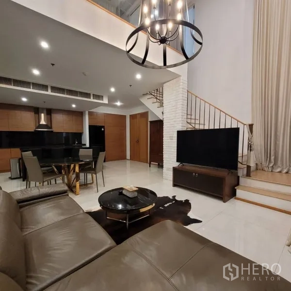 Condominium for rent in Khlong Toei, Bangkok - Lounge with large TV, staircase to mezzanine, and sleek kitchen in the background.