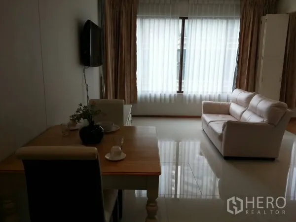 Condominium for rent in Khlong Toei, Bangkok - Bright living area with large windows, white sofa, curtains, and wall-mounted TV beside a dining nook.