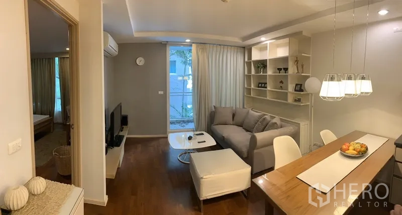 Condominium for rent or sale in Khlong Toei, Bangkok - Bright lounge with TV console, sliding glass door to a leafy balcony, feature shelving, and a dining table under pendant lighting.