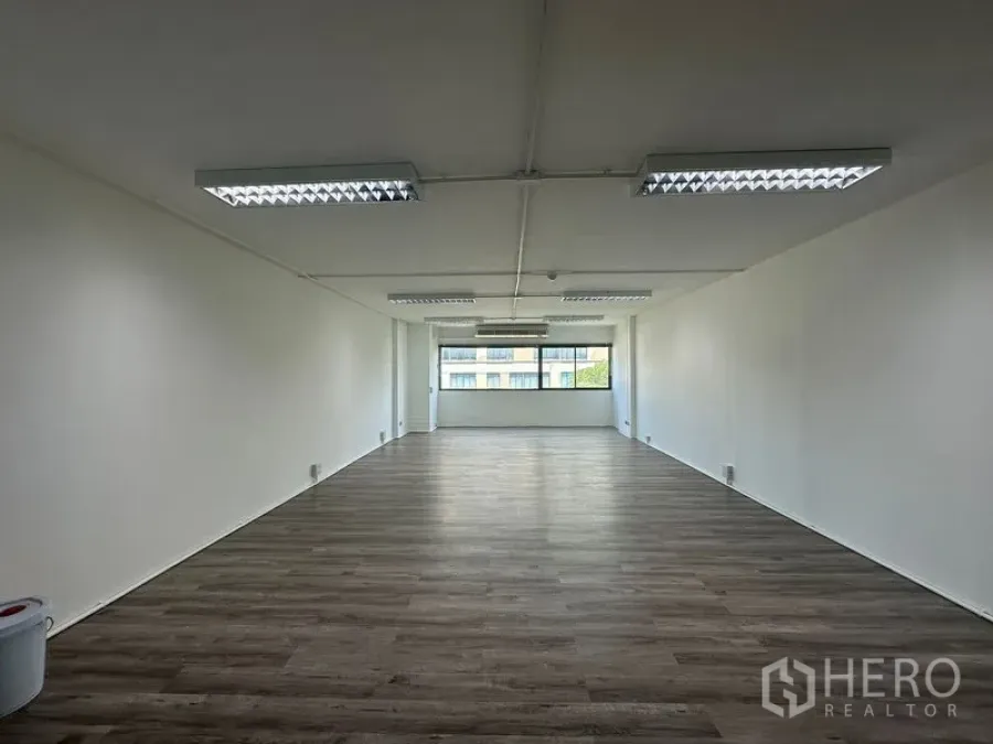 Office Space for rent in Bangkok - Empty office space with ceiling lights, white walls and a windowed facade overlooking nearby buildings.