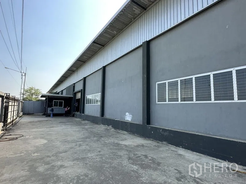 Factory for rent in Lam Luk Ka, Pathum Thani - Long exterior wall with multiple windows and broad concrete apron for truck access.