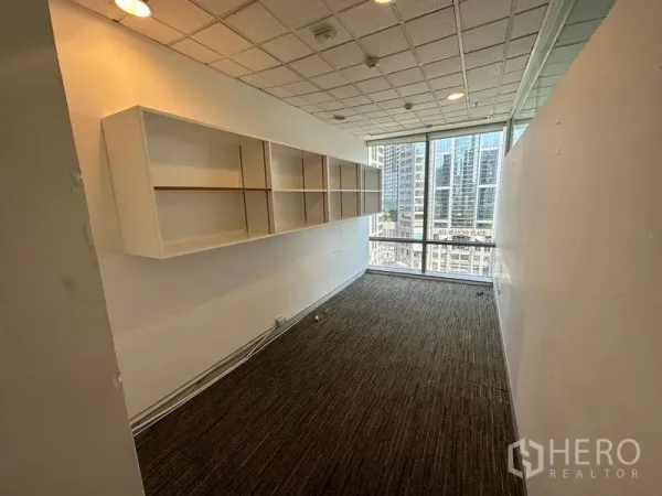 Office Space for rent - Narrow office room with built-in white shelving, carpet flooring and a floor-to-ceiling window facing the Bangkok skyline.
