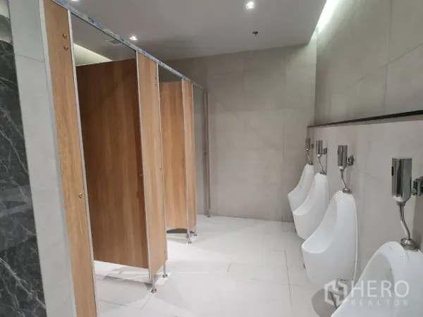 Office Space for rent - Washroom view showing wooden cubicle doors and wall-mounted urinals with light tiled walls and floor.