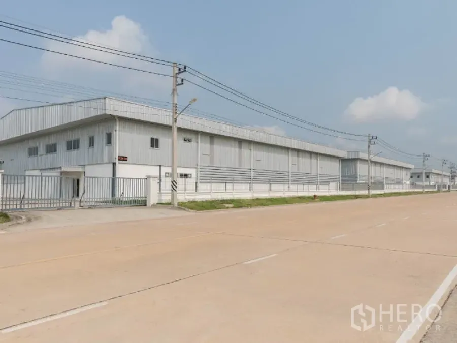 Factory for rent in Si Racha, Chon Buri - Long metal-clad factory exterior along a wide industrial road with gated frontage and power lines.