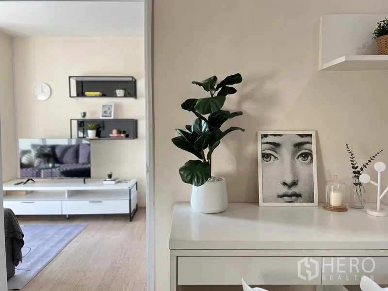 Condominium for rent in Khlong Toei, Bangkok - Minimalist condo nook with potted plant and decor, opening to a bright living room with TV console and wall shelves.