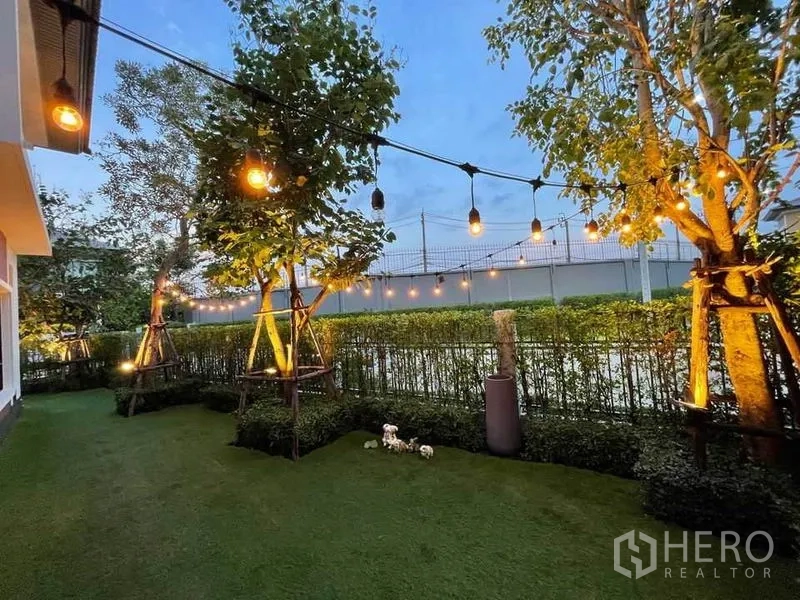 Detached House for sale in Pak Kret, Nonthaburi - Backyard lawn with string lights, trees and trimmed hedges at twilight.