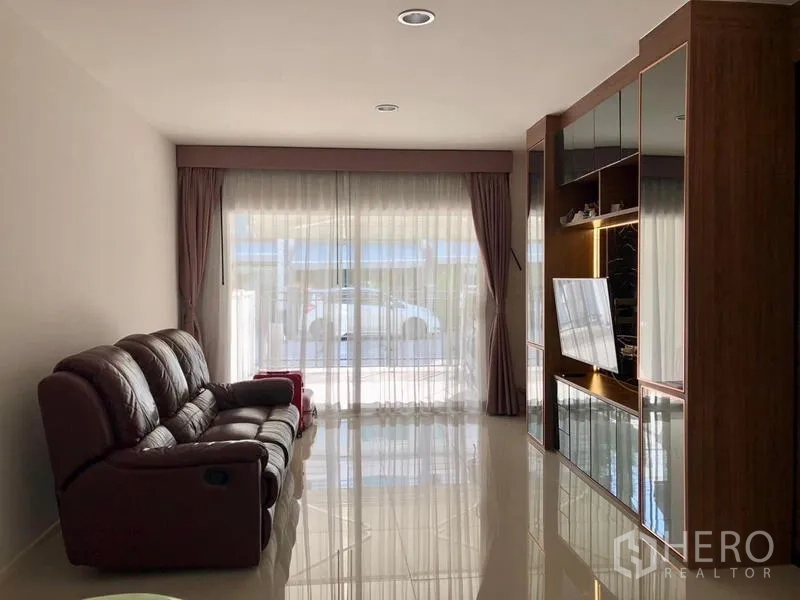 Townhouse for rent in Suan Luang, Bangkok - Sunlit living room with leather sofa, floor-to-ceiling sliding glass doors and a built-in TV wall unit.