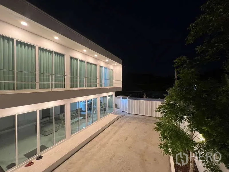 独立式住宅 出租在Lat Krabang, Bangkok - 夜色下的庭院与玻璃立面两层住宅，灯光通明。