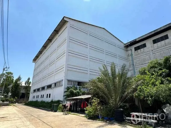 工厂 出租在Mueang Samut Prakan, Samut Prakan - 大型白色工业建筑的背侧，沿小路有绿化植物。