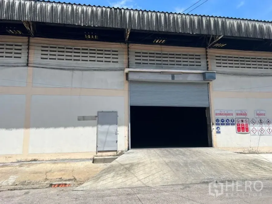Warehouse for rent in Mueang Samut Prakan, Samut Prakan - Warehouse facade with ramped loading dock, roller shutter entrance and safety signage.