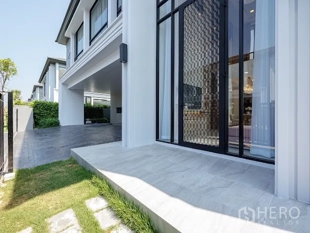 Detached House for sale in Prawet, Bangkok - Side terrace and tall feature window beside covered carport and trimmed lawn.