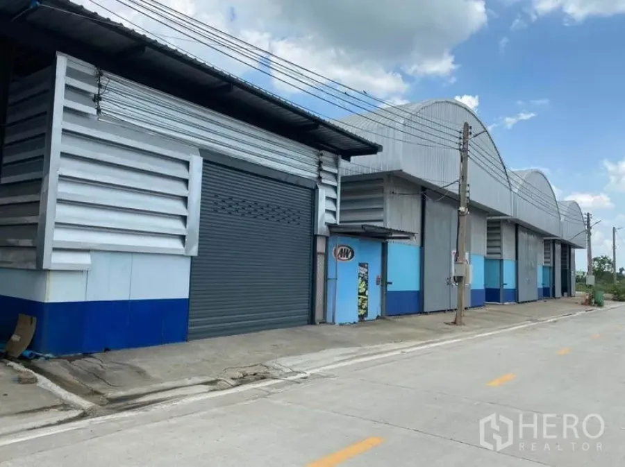 Warehouse for rent in Khlong Luang, Pathum Thani - Row of metal-clad warehouses with roller shutter doors along a wide concrete road under blue sky.