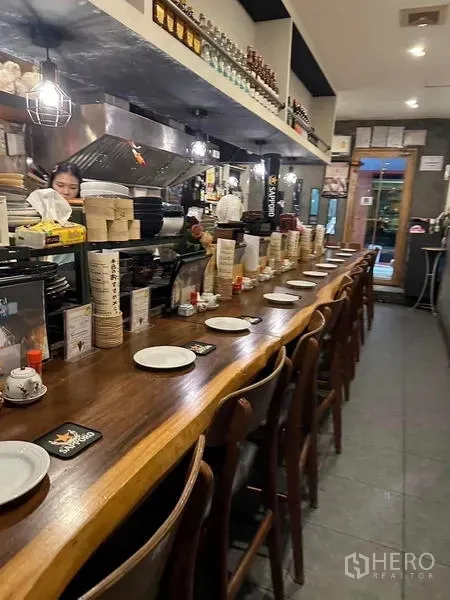 Shophouse for rent or sale in Watthana, Bangkok - Long wooden bar counter with rows of chairs and table settings leading toward the entrance.