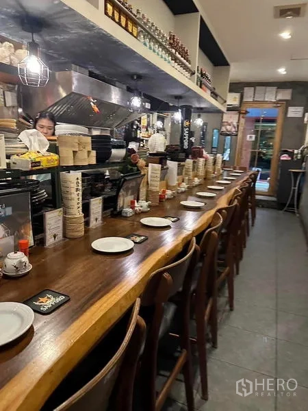 Shophouse for rent or sale in Watthana, Bangkok - Long wooden bar counter with rows of chairs and table settings leading toward the entrance.