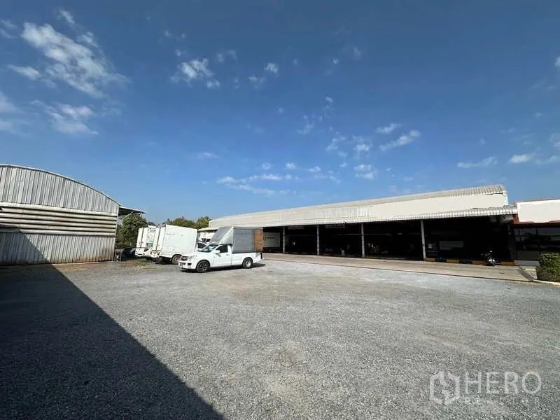 Warehouse for rent in Lam Luk Ka, Pathum Thani - Gravel parking court opposite a long shed and part of the main warehouse.