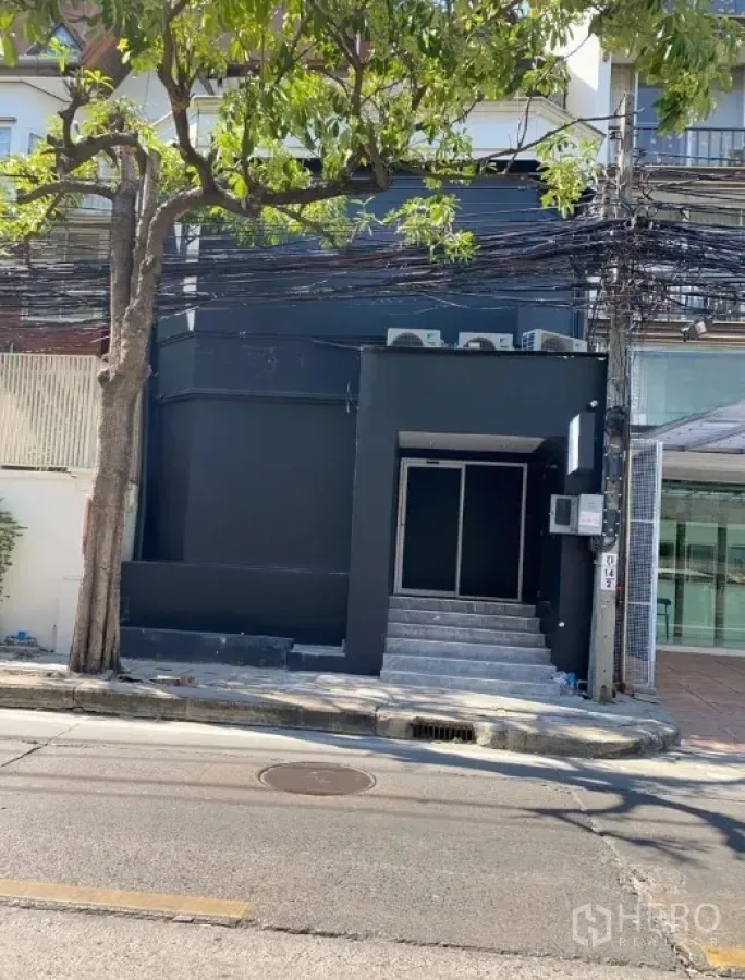 店屋 出租在Bangkok - 黑色外立面的商铺门面，台阶入口，街旁绿树与电线。