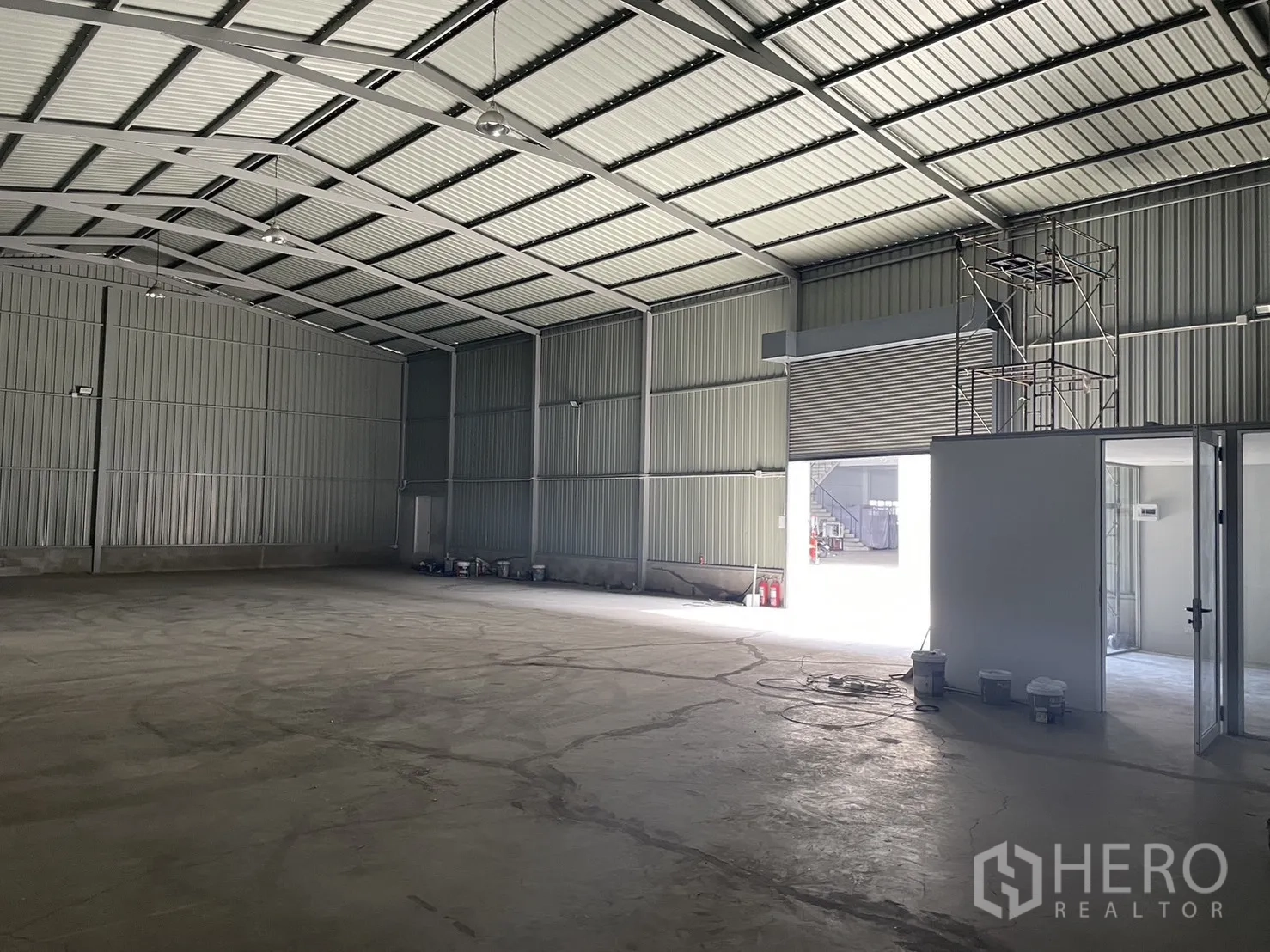 Factory for rent in Mueang Samut Prakan, Samut Prakan - Spacious steel-frame interior with high ceiling, concrete floor, and roller shutter opening with a small room.