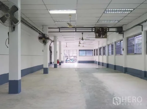 Warehouse for rent in Bang Kapi, Bangkok - Spacious hall with concrete floor, wall-mounted fans and barred windows providing airflow.