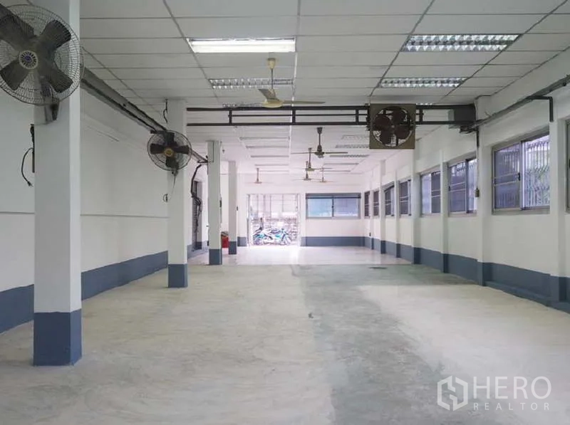 Warehouse for rent in Bang Kapi, Bangkok - Spacious hall with concrete floor, wall-mounted fans and barred windows providing airflow.