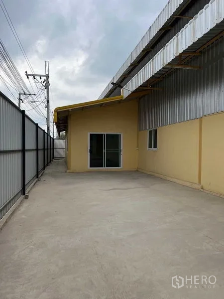 Warehouse for rent in Lat Krabang, Bangkok - Concrete driveway and security gate beside a metal‑clad warehouse with a sliding glass entry.