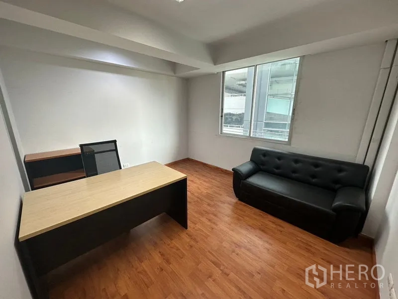 Office Space for rent in Khlong Toei, Bangkok - Private office with wood-look flooring, a modern desk and chair, black leather sofa, and a window bringing in natural light.