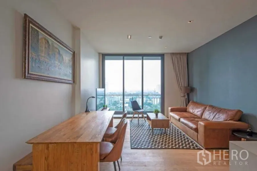 Condominium for rent in Khlong Toei, Bangkok - Open-plan living and dining area with leather sofa, long wooden table, large glass wall and expansive city view.